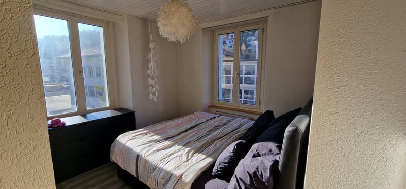 Bedroom with bed, pillows, and a nightstand with a lamp. Two windows with a view of the outside.