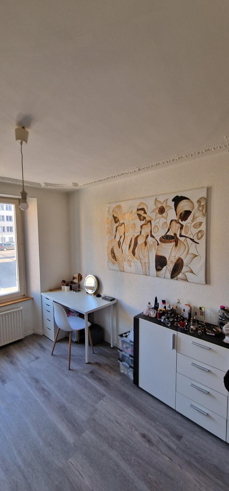 A room with a desk, chair, and cabinet. A painting is on the wall. A radiator is on the left side of the room. A window is on the left side of the room. A lamp is hanging from the ceiling.