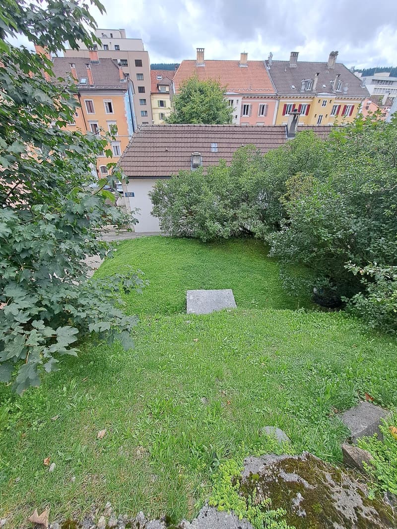 A small garden with green grass, a few trees, and a stone slab in the middle. The garden is surrounded by buildings and trees.