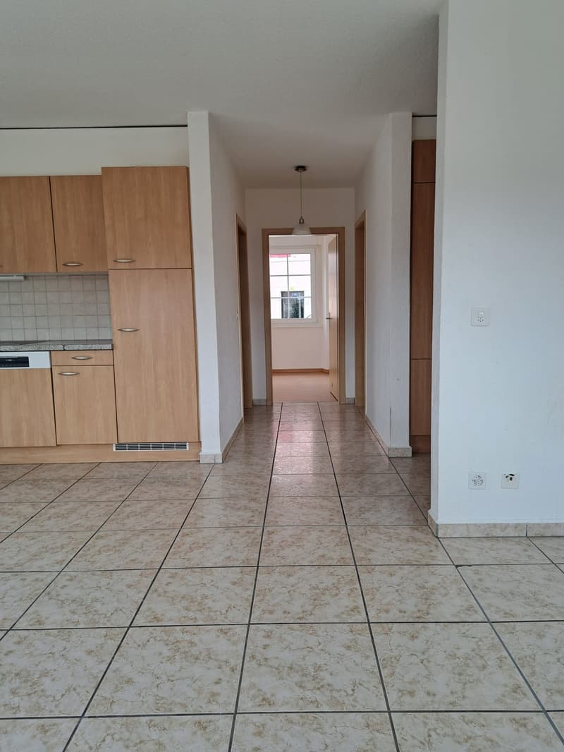 Empty hallway with tiled floor, wooden cabinets, kitchen area with dishwasher, and doors leading to other rooms