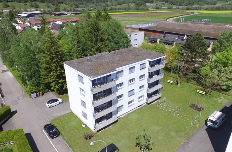 A multi-story apartment building with balconies, surrounded by trees and grass, with a playground and parking lot in front.