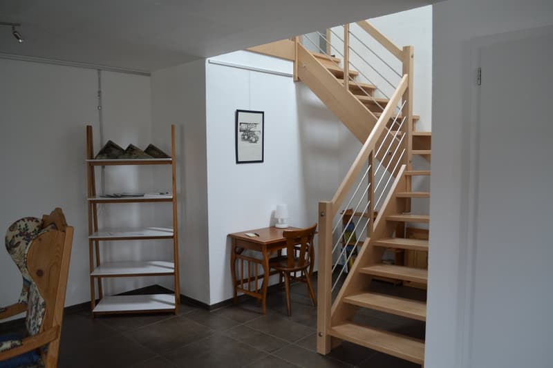 The image shows the interior of a room with a wooden staircase leading to an upper level. There is a wooden shelf unit on the left side of the room, and a small wooden table with two chairs in the center. The walls are white, and the floor is tiled. The s