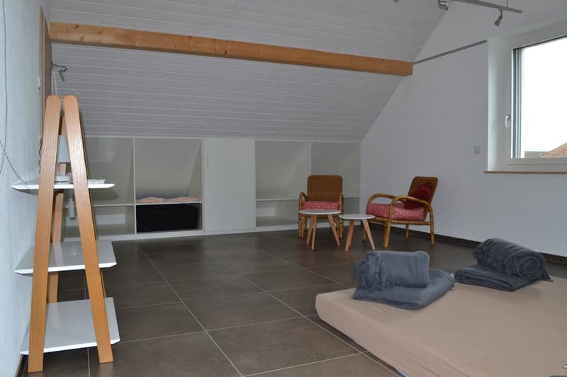 Attic room with slanted ceiling, wooden beams, gray tiled floor, built-in shelving units, two armchairs, and a sofa with blankets