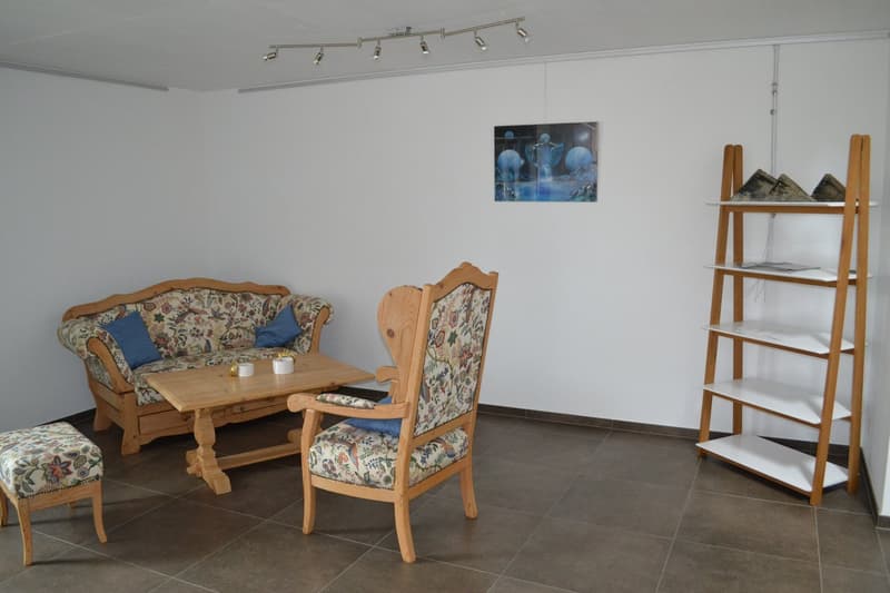 Cozy living room with floral patterned sofa and armchair, wooden coffee table, and a shelving unit. The room has a tiled floor and track lighting on the ceiling.
