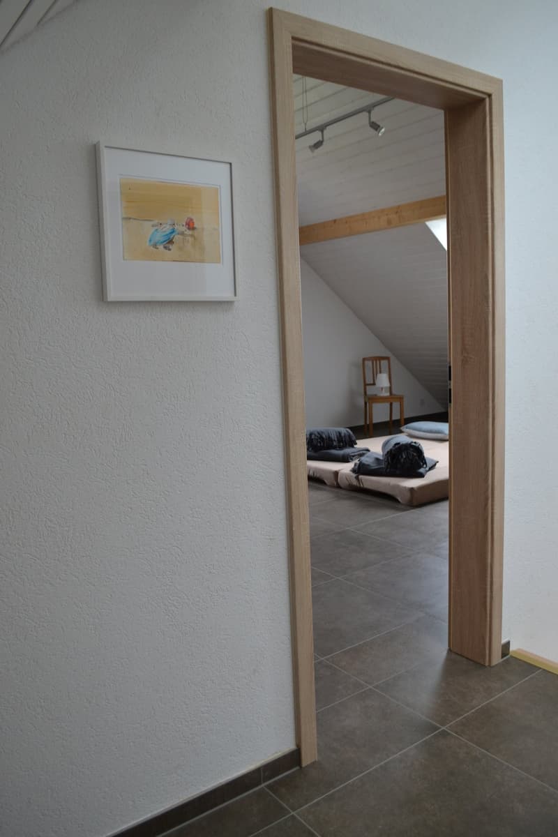 The image shows a room with a wooden framed mirror on the wall, a framed artwork, and a tiled floor. Through the doorway, a bedroom can be seen with a bed and chairs.