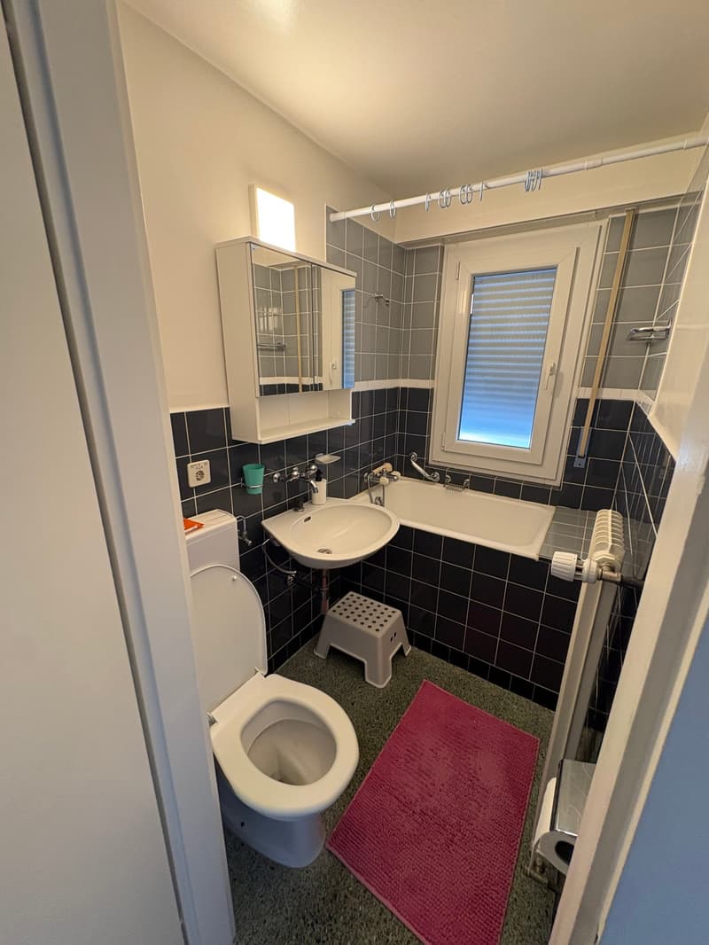 Bathroom with toilet, sink, bathtub, window, cabinet, and tiled flooring.