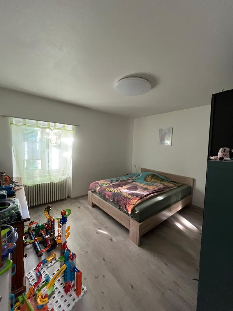 A cozy bedroom with a wooden bed, a desk with toys, a radiator, a window with curtains, a picture frame on the wall, and a green closet