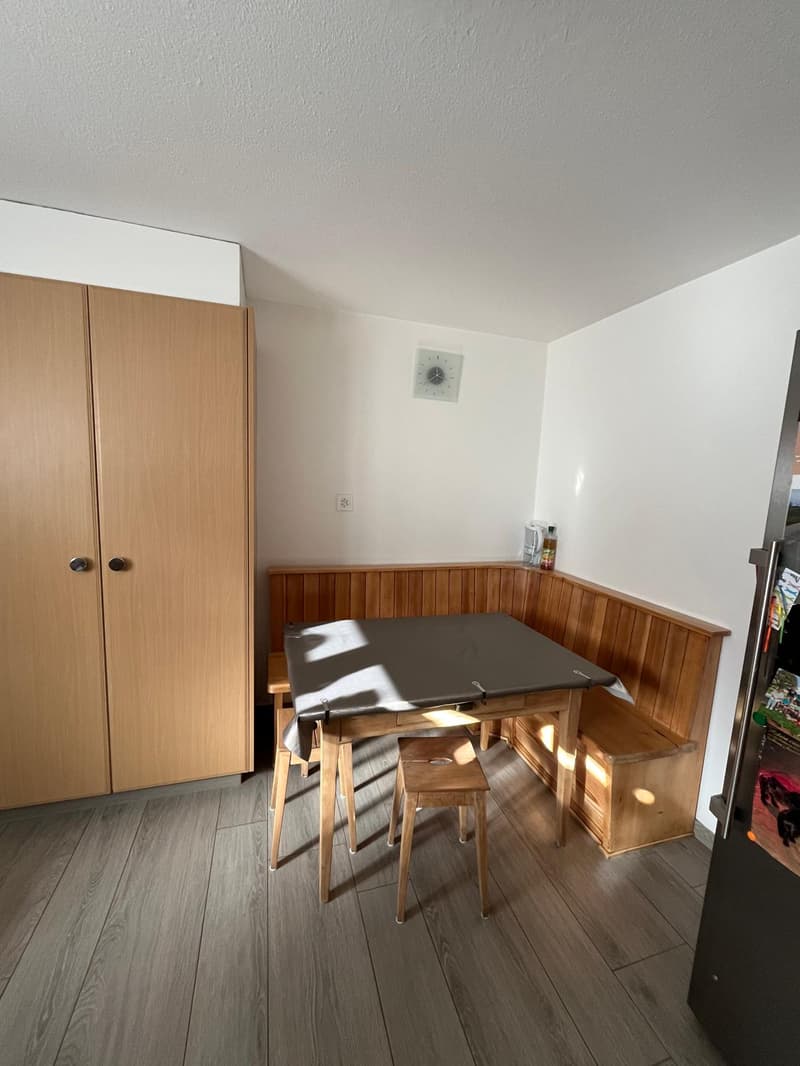 Wooden desk, wooden stools, wooden dining area, wooden cupboard, wall clock, power outlet, and a white wall