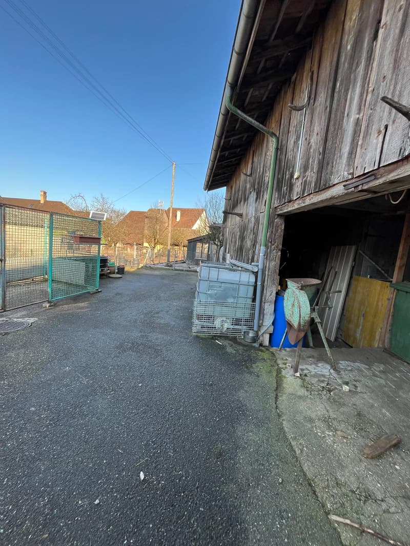 Wide entrance road leading to an old building, a fence, storage items, and a green gate.
