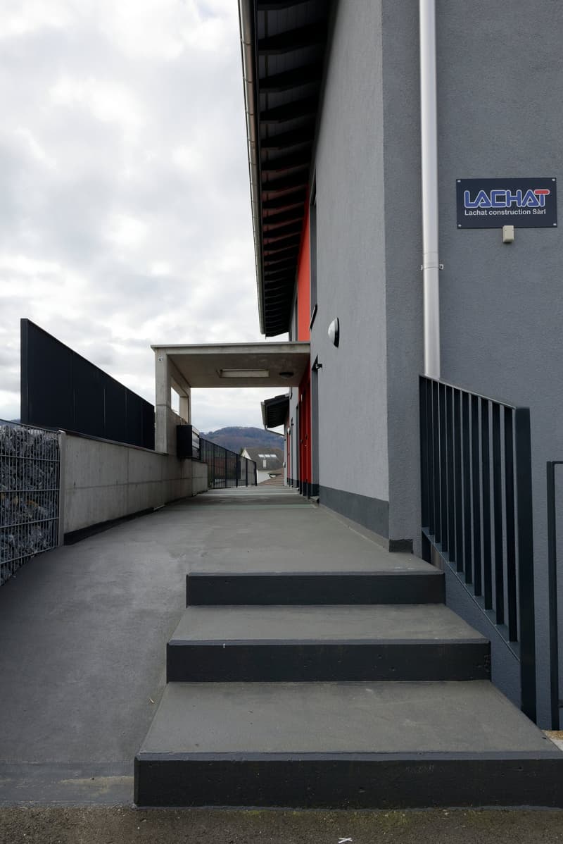 Exterior of a building with a raised ground floor, concrete steps, metal railings, and a sign reading 'LACHAT construction Sàrl'.