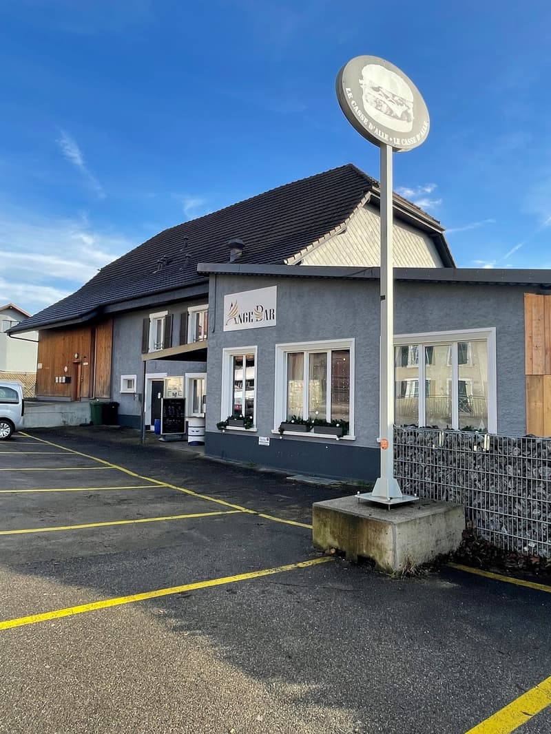 grey facade with wooden accents, multiple windows, trash bins, sign, paved parking lot, stone fence