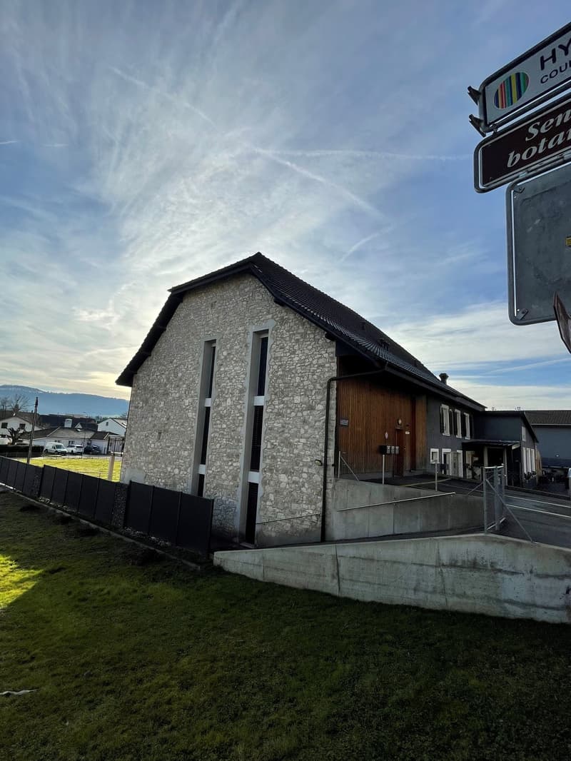 A stone exterior two story house, gray roof, large windows, wooden door, small parking area, black fence, grassy yard.