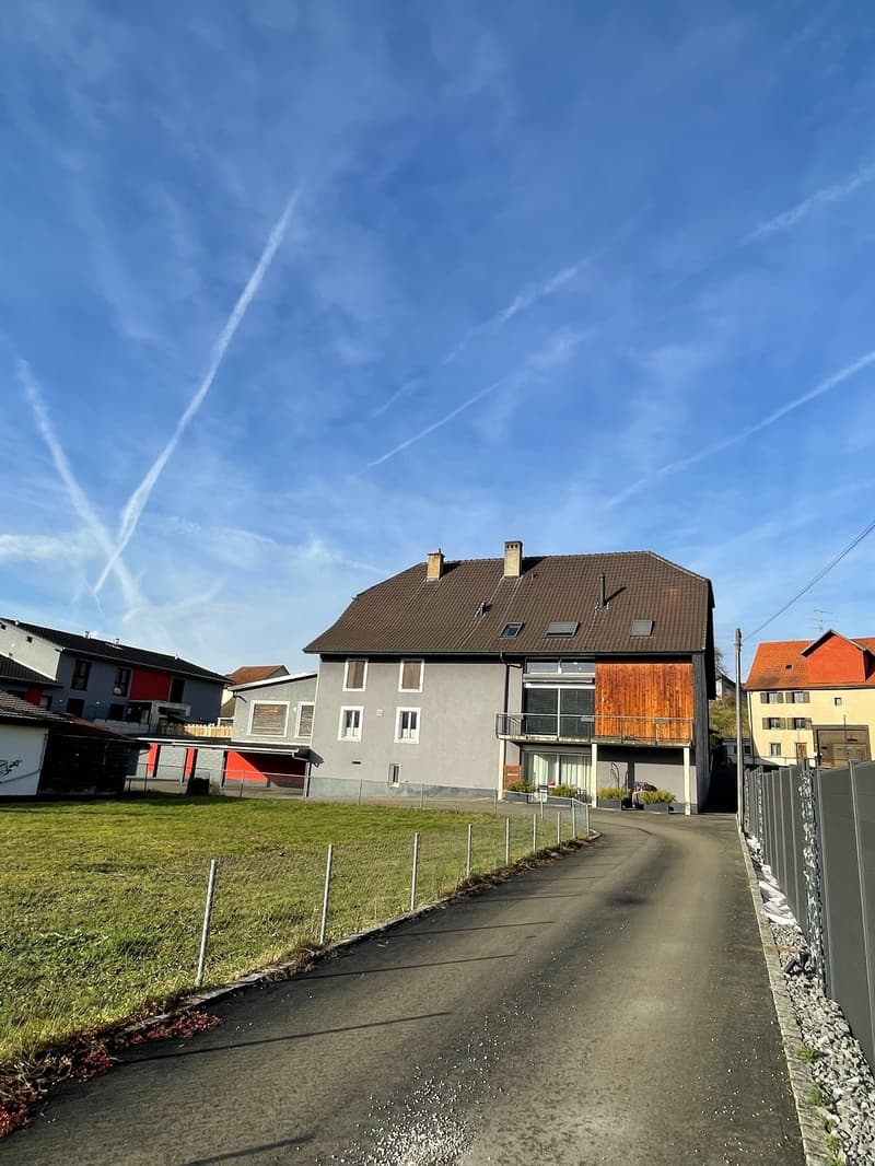 gray building, brown roof, brown wood paneling, balcony, large windows, fenced yard, driveway, grassy lawn