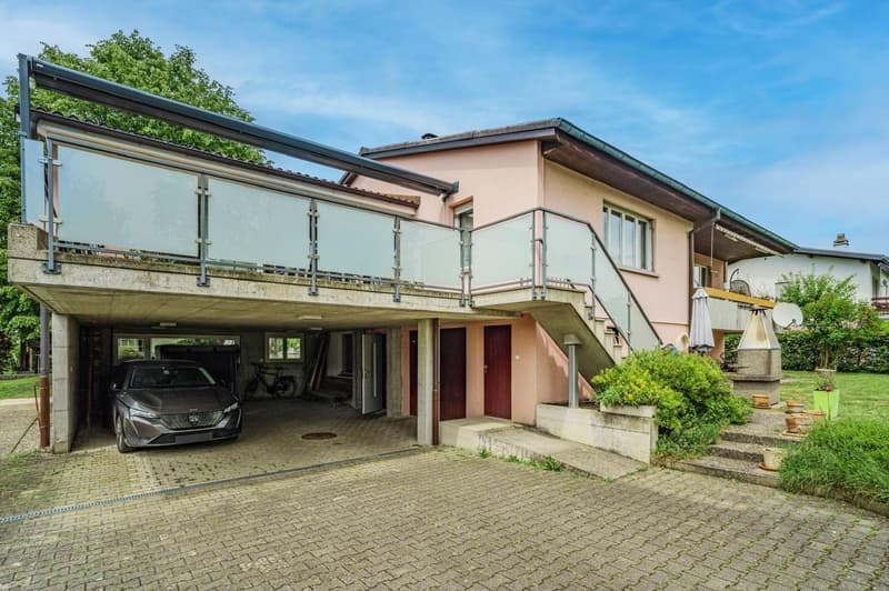 2-story house with a balcony, garage, and parking space. The house has a pink exterior with large windows and a tiled roof. There are plants and greenery surrounding the property.