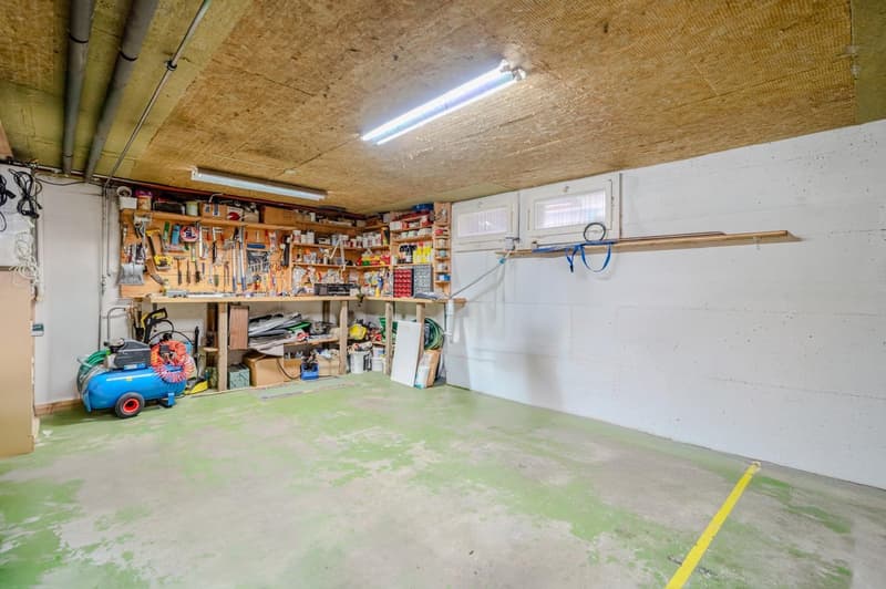 Cluttered garage space with various tools and equipment stored on shelves, a blue air compressor, and a concrete floor with yellow markings.
