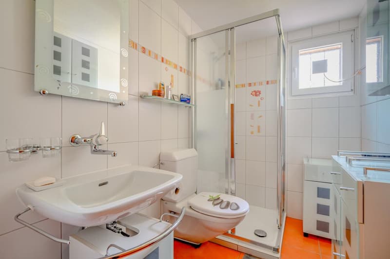 Tiled bathroom with a sink, toilet, and shower enclosure. The room has white walls and floors with orange accents. There is a washing machine and dishwasher visible in the room.