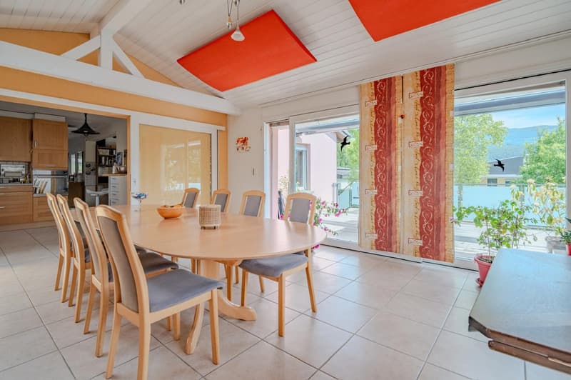Spacious dining area with large windows overlooking a scenic outdoor view, wooden dining table and chairs, orange accent ceiling panels, and patterned curtains.