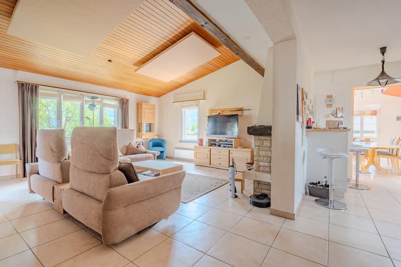 Open-concept living room with vaulted wood ceiling, large windows, beige sofa and armchair, TV stand, and kitchen area visible in the background