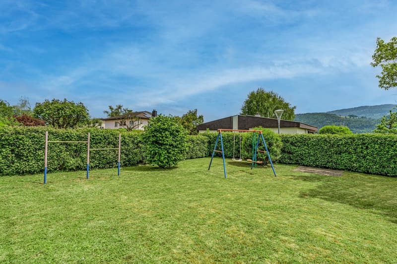 2-story house with a well-maintained garden, including a grassy lawn, hedges, and a children's playground with swings. The house has a tiled roof and is surrounded by trees and greenery.