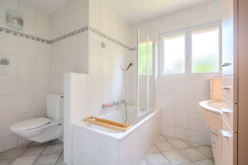 Spacious bathroom with white tiled walls, a bathtub, a shower enclosure, a toilet, and a vanity unit with a sink. The room has large windows providing natural light.