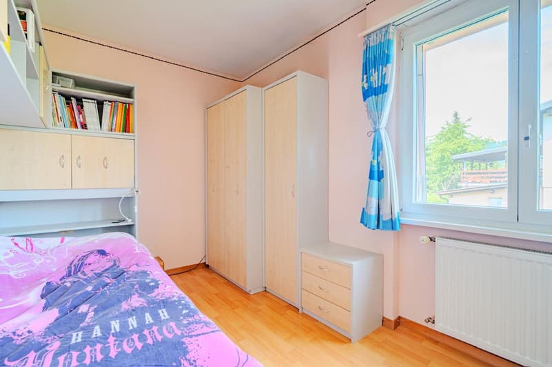 Bedroom with pink and blue color scheme, wooden floor, built-in shelves, wardrobe, dresser, and large window with blue patterned curtains