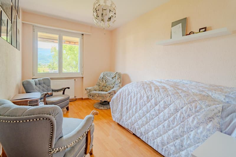 Spacious bedroom with large windows, hardwood floors, a king-size bed with a quilted comforter, two upholstered armchairs, and a decorative chandelier.