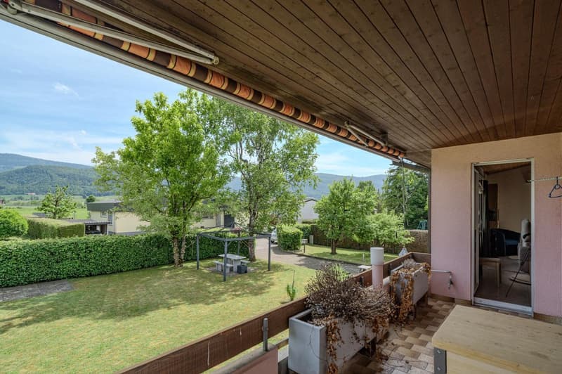 Covered patio or balcony with wooden ceiling, overlooking a lush green yard with trees and shrubs, surrounded by mountains in the background