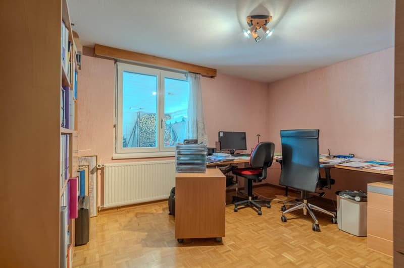 The image shows a home office space with a desk, computer, bookshelves, and other office equipment. The room has wooden floors, pink walls, and a window overlooking what appears to be a backyard or outdoor area.