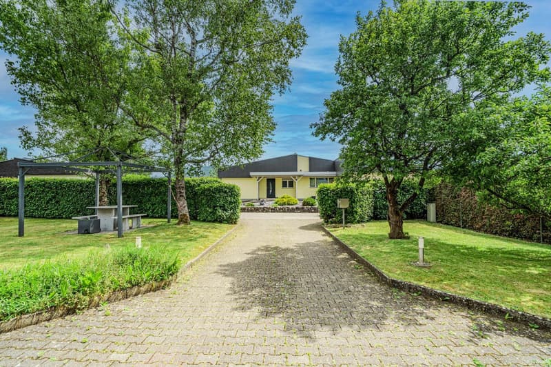 Single-story yellow house with a paved walkway leading to the entrance, surrounded by lush green trees and a well-manicured lawn. There appears to be a covered patio or pergola structure in the yard, and a parking area is visible as well.