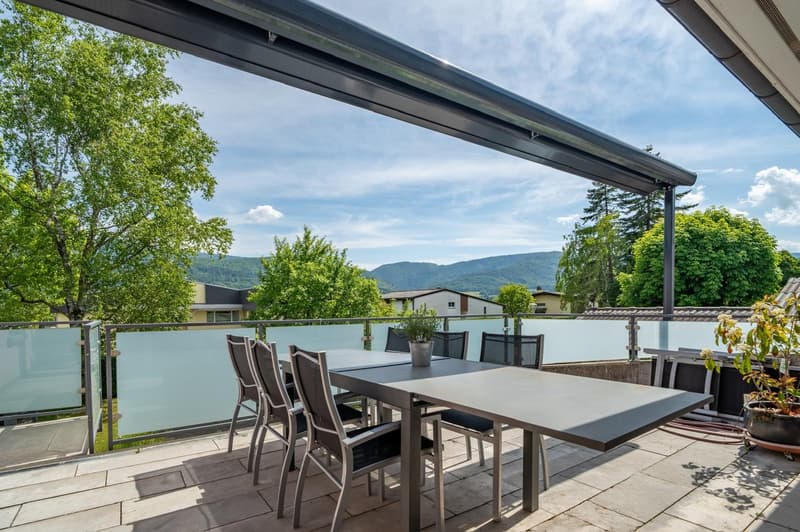Covered outdoor patio with a dining table and chairs, overlooking a scenic view of trees and mountains in the background.
