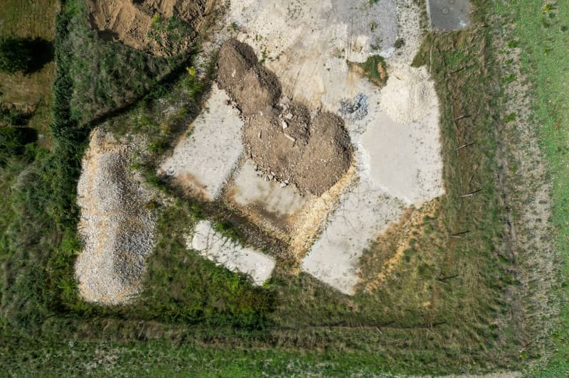 The image shows an aerial view of a natural landscape. It appears to be a forested area with a clearing or damaged section in the center. The ground is covered in dirt, rocks, and vegetation, with some areas of exposed soil or mud. The surrounding area is