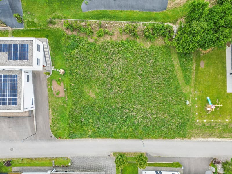 This image shows an aerial view of a building with solar panels on the roof, surrounded by a large grassy area with some trees and a paved parking area.