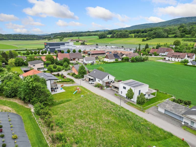 This image shows an aerial view of a small rural town or village surrounded by lush green fields and hills. The town consists of a mix of residential houses, some larger modern buildings, and a few commercial or industrial structures. There is a parking a