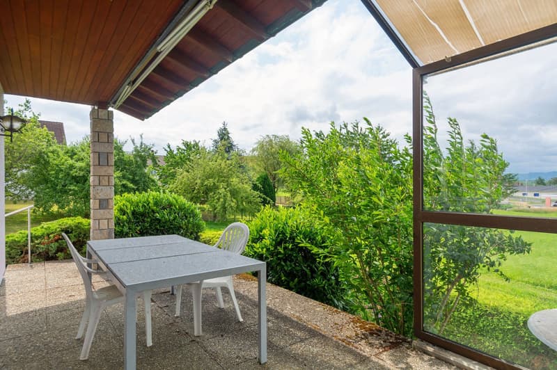 Covered patio with a table and chairs, surrounded by lush greenery and trees. The patio has a wooden roof and is connected to the main building.