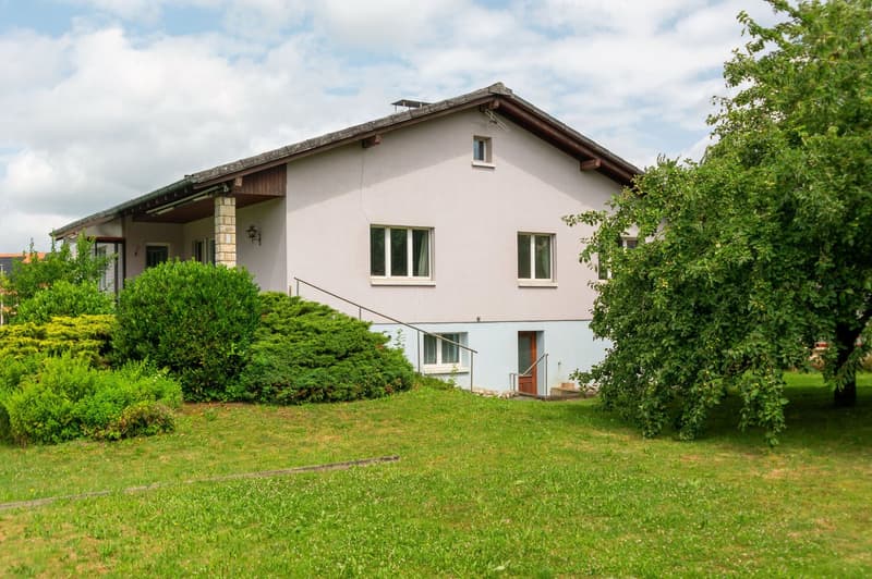2-story house with a white exterior, wooden roof, and large windows. The house is surrounded by a well-maintained lawn and lush greenery, including trees and shrubs. There appears to be a balcony or terrace on the upper level of the house.