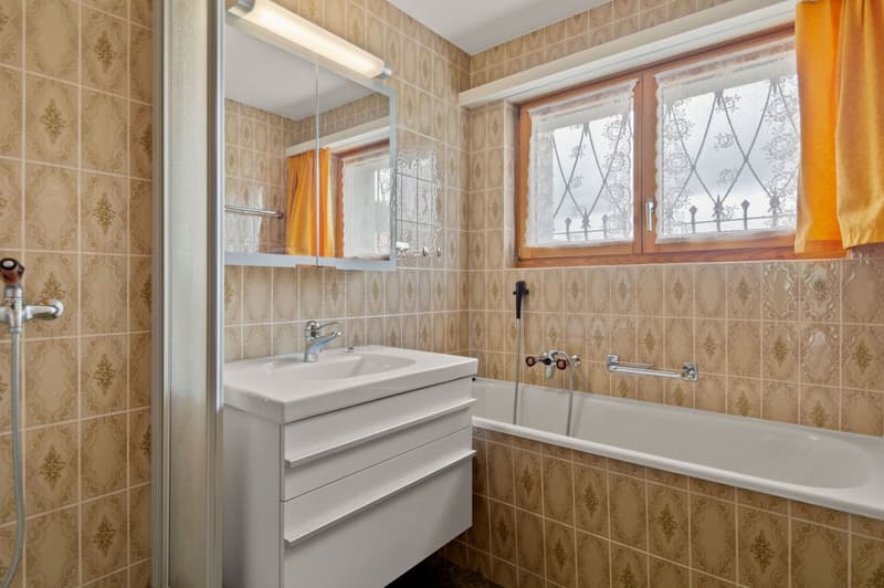 The image shows a bathroom with beige patterned tiles on the walls, a white vanity with a sink, a bathtub, and a mirror above the vanity. The bathroom also has a window with decorative curtains and a shower enclosure.