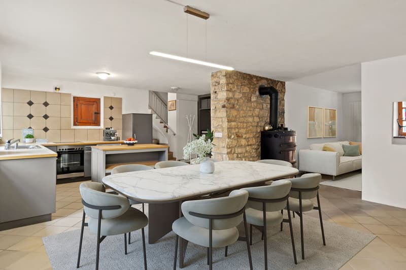 Open plan kitchen, modern appliances, marble countertop, large dining table, integrated seating area, decorative wall tiles, large window, gray and white decor.