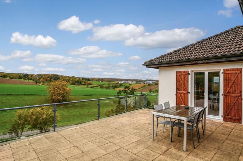 Outdoor dining area with table and chairs, metal railing, view of green field, modern white walls, wooden shutters