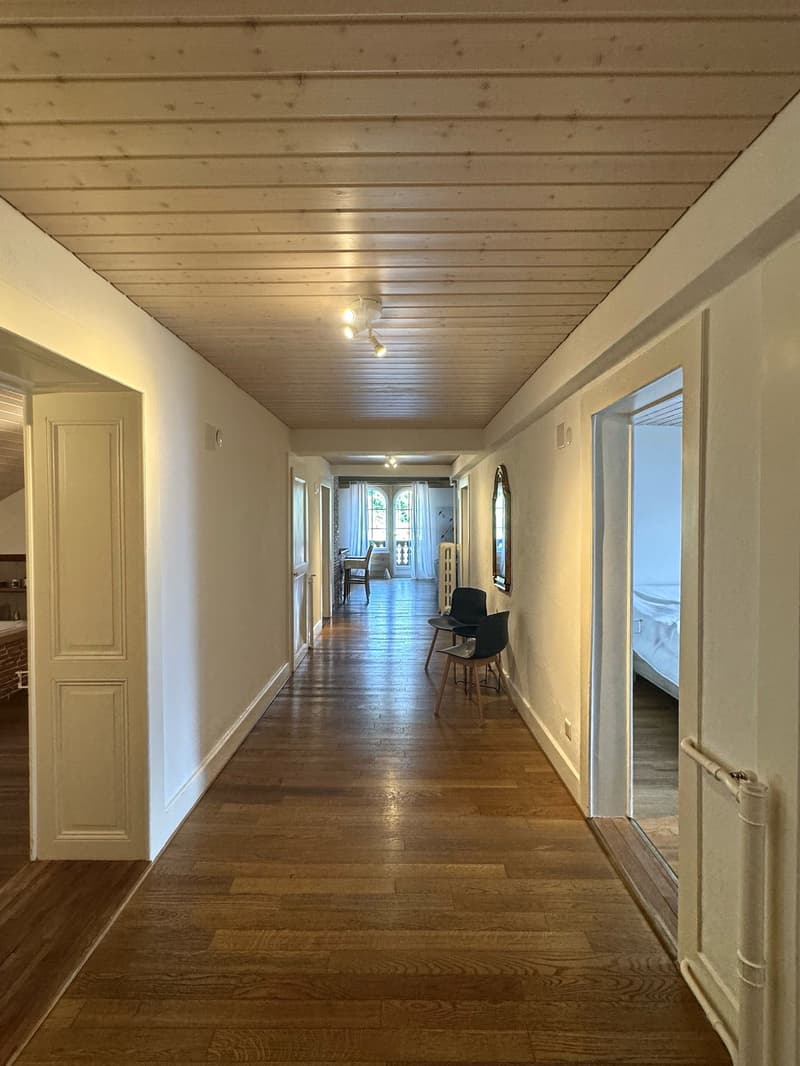 An empty hallway with hardwood floors and white walls. On the left side, there are two doors, one of which leads to a bathroom with a bathtub and a mirror. On the right side, there are two open doors, one leading to a bedroom with a bed, and the other lea