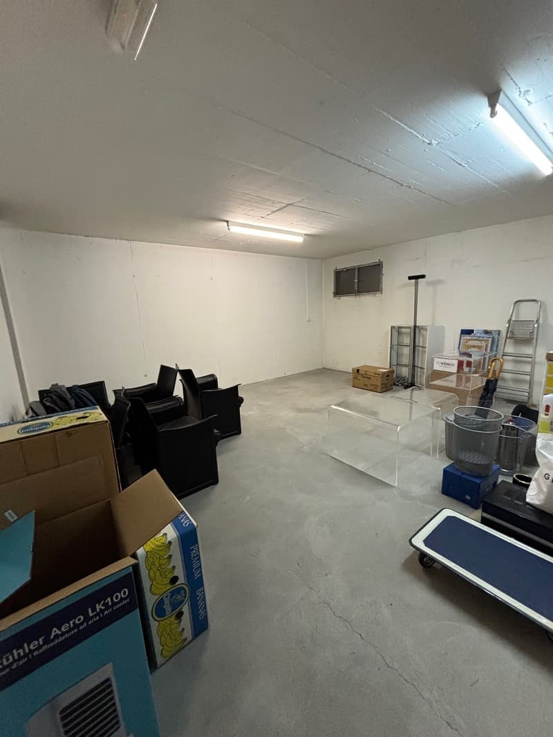Empty room with white walls, concrete floor, ceiling lights, storage boxes, chairs, ladder, and other items