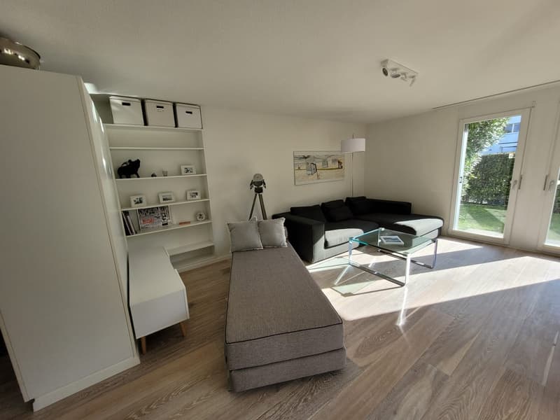 modern living room with gray sofa, glass table, wooden floor, white shelves, and sliding glass doors leading to a garden