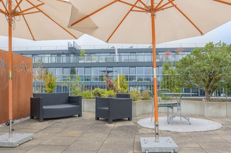 Outdoor seating area with two large umbrellas, a table, and two chairs, near a metal wall, and a staircase leading up to an apartment building in the background.