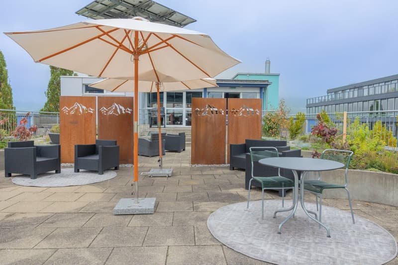 covered seating area with tables, chairs, and umbrellas, stone floor, fenced garden, view of buildings in the background
