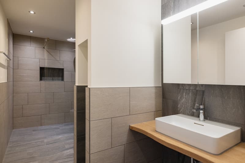 modern shower area with glass wall, tiled flooring, wooden countertop, white sink with silver faucet, mirror with light