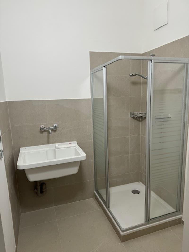 The image shows a bathroom with a glass shower enclosure, a wall-mounted sink, and beige tile walls.