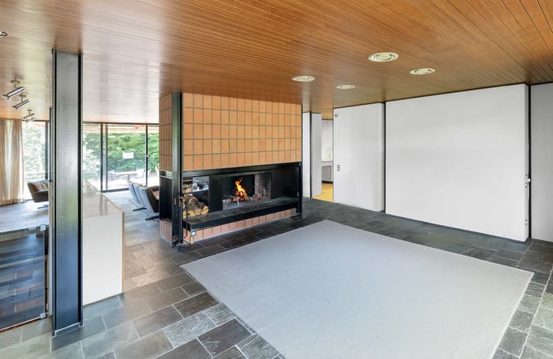 wooden ceiling, fireplace, black fireplace hearth, white rug, white walls, large windows, glass wall, chairs, brown tile floor