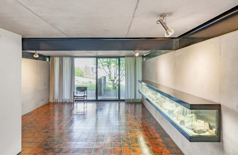 Modern room with red tile flooring, large sliding glass door leading to a balcony, white walls, track lighting, a chair, and a built-in display shelf with an aquarium.
