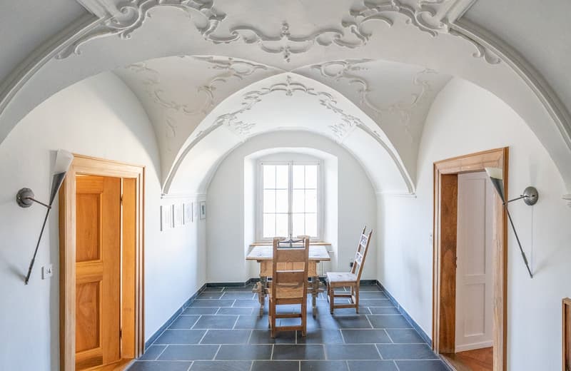 This image shows a vaulted ceiling room with ornate, decorative plasterwork. The room has a wooden table and chairs in the center, and the floor is covered with blue slate tiles. There are wooden doors on either side of the room, and a large arched window