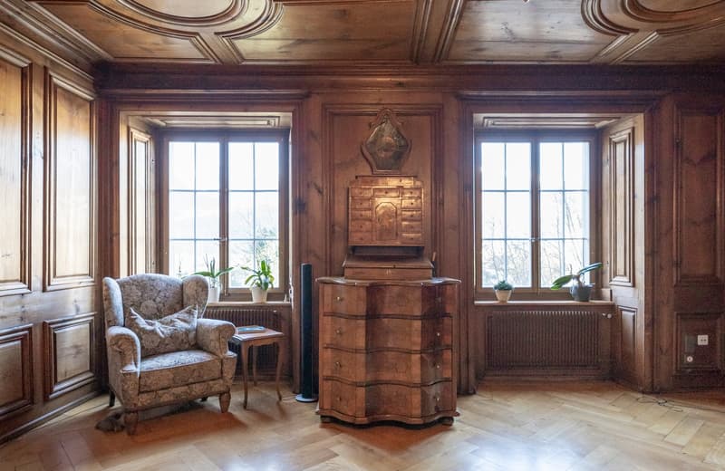 Ornate wooden paneled walls and ceiling, large windows, antique wooden dresser, armchair, potted plants