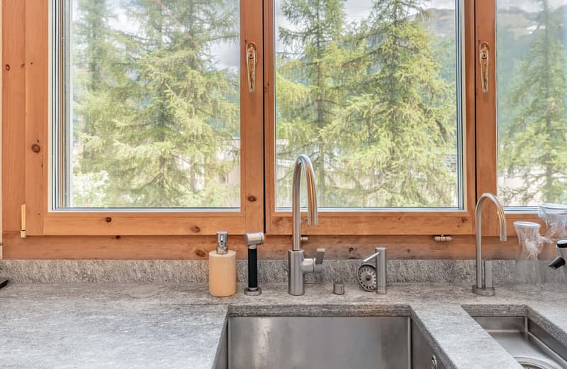 Spacious kitchen with large windows overlooking a forested area, granite countertops, stainless steel sink and faucet, wooden cabinetry and trim